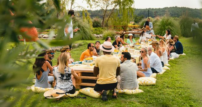 A grand celebration with a large gathering of people seated around a table, nestled on pillows in the park.